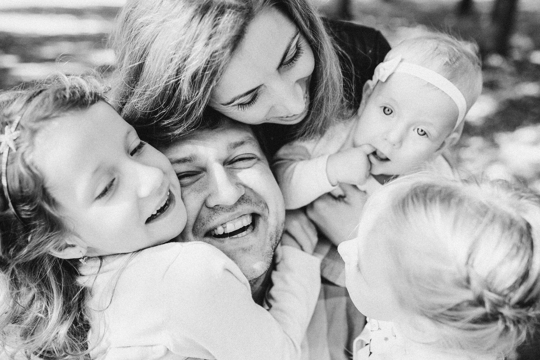 Black-and-white photo of a smiling family of four in a tight hug outdoors, showing dad, mom, and two young children.