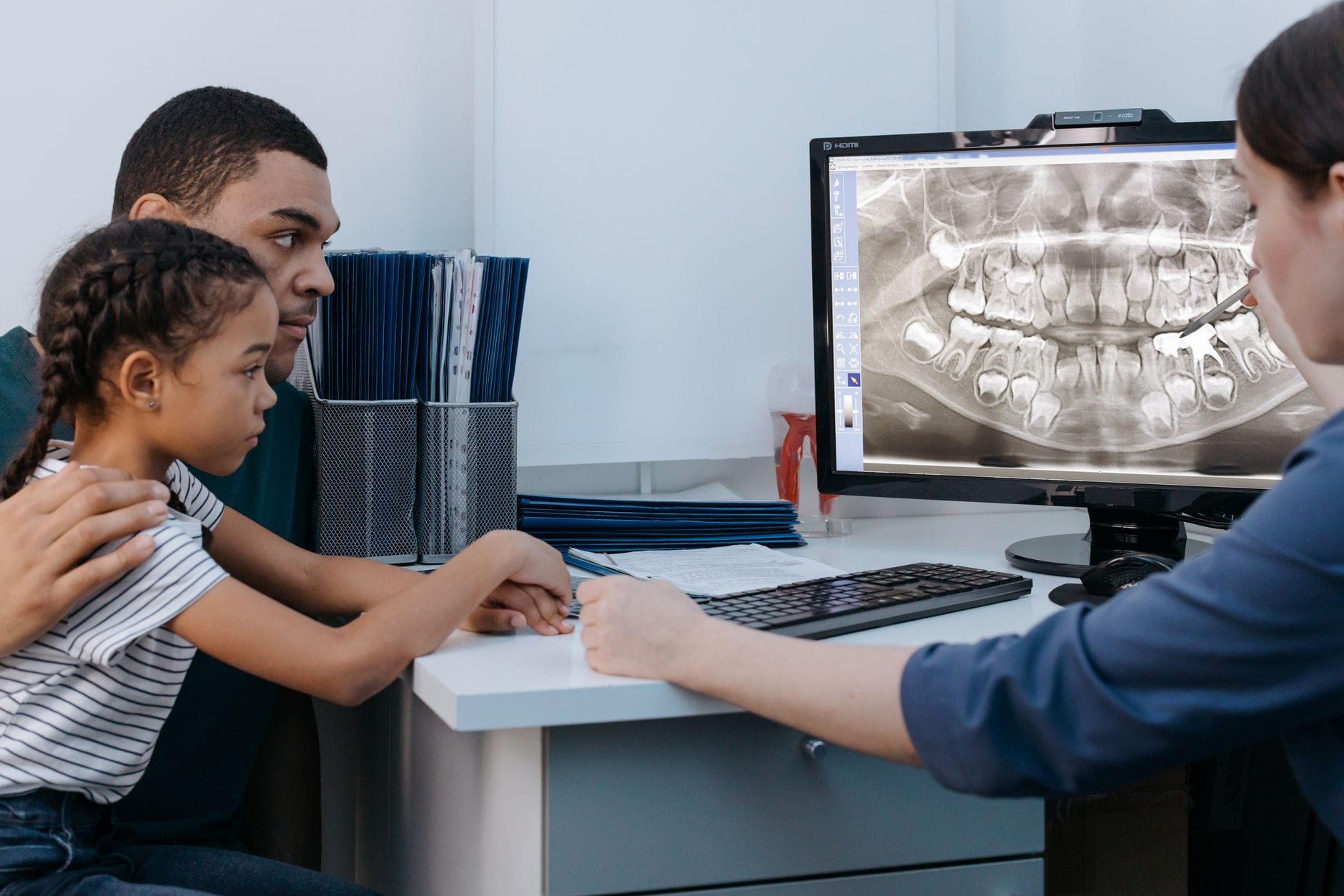 Dentist explains a dental X-ray on a computer screen to a young patient and parent at a desk.