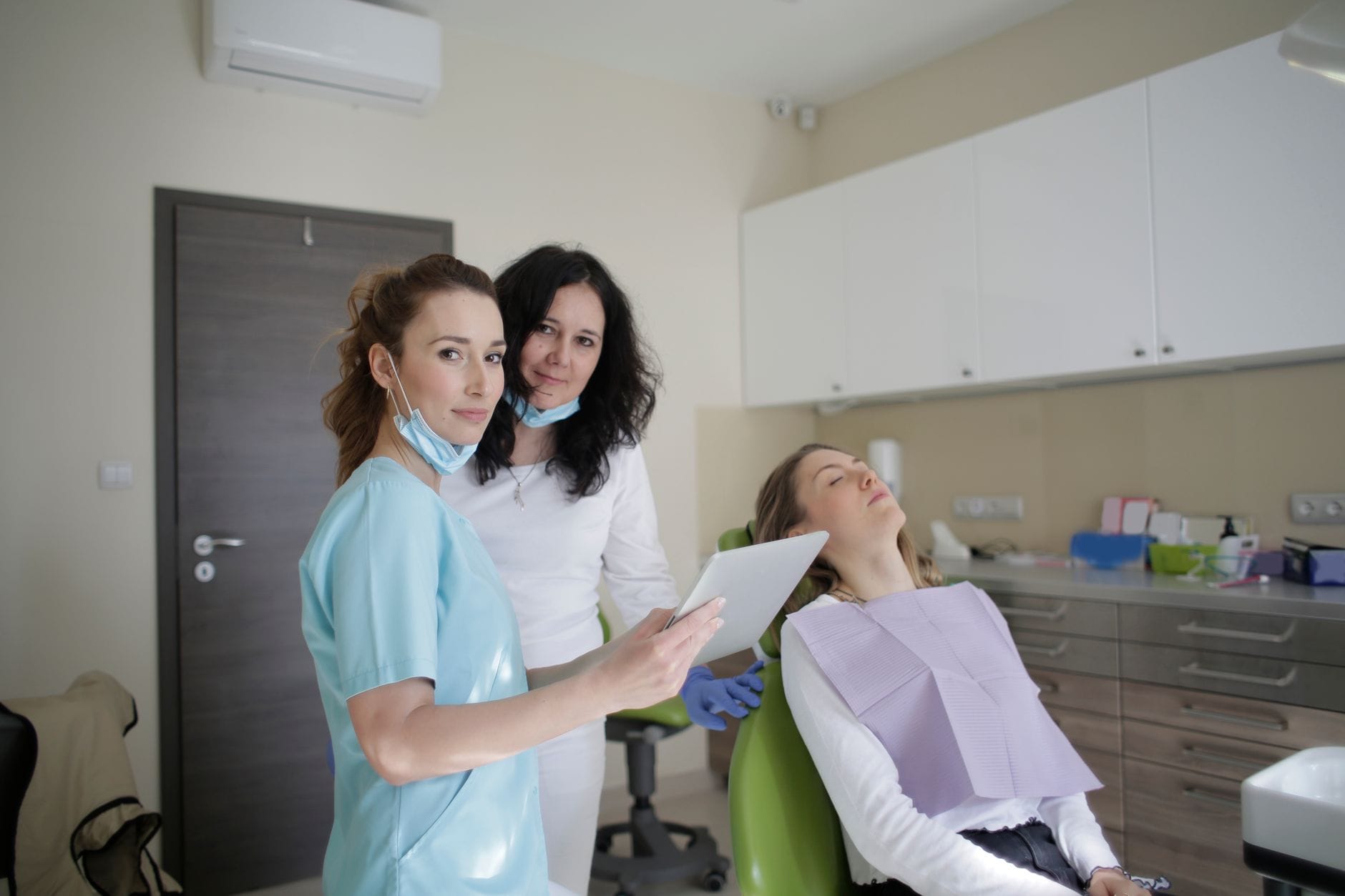 Dentist in blue scrubs and two assistants stand beside a patient reclining in a dental chair in a modern clinic.