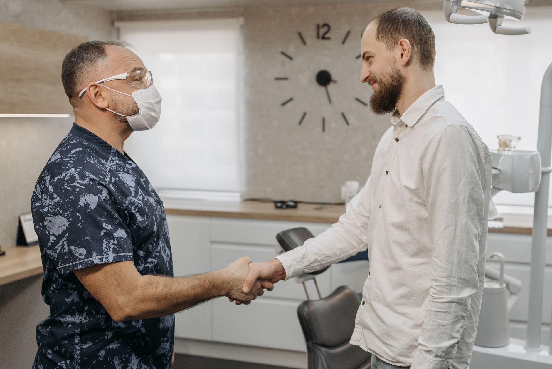 Two men shake hands in a dental clinic; one wears a face mask and dark camo-pattern scrubs, the other in a light shirt, smiling.