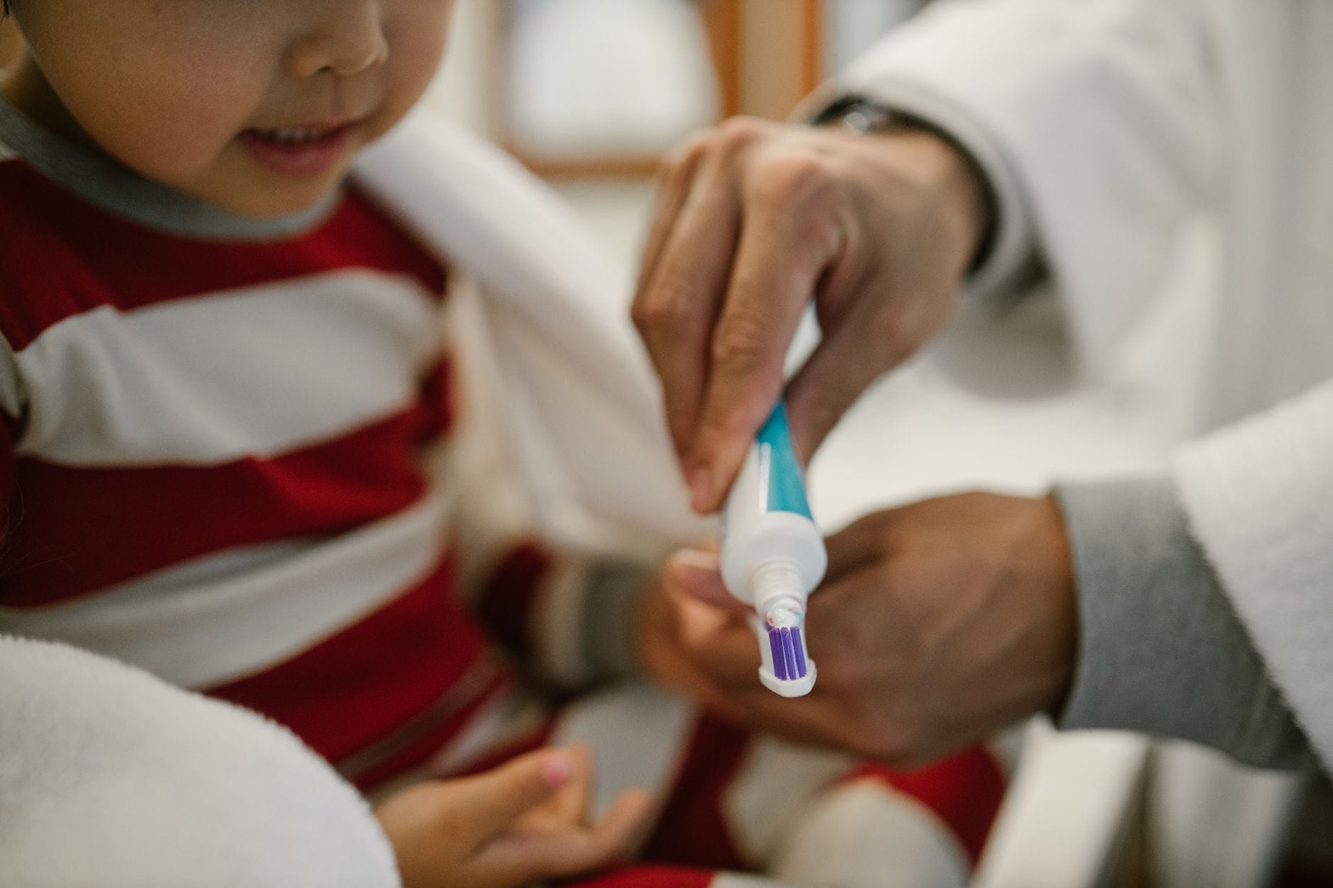 A young child in a red-and-white striped outfit having their teeth brushed by a caregiver using a toothbrush.
